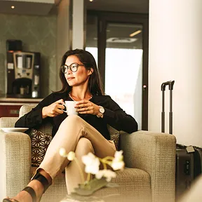 A businesswoman relaxes in a premium lounge, enjoying coffee beside her suitcase in a calm, elegant setting.