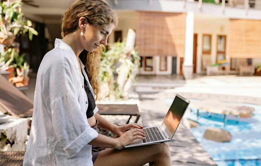 A woman works remotely on her laptop beside a tranquil pool, enjoying a sunny outdoor setting at a modern villa.